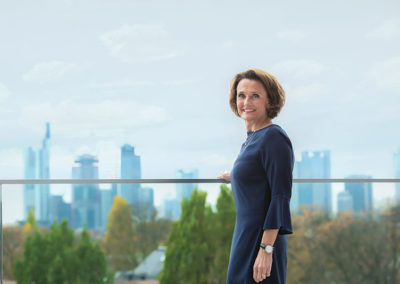 Auf dem Bild ist Helen Hain zu sehen. Sie trägt ein dunkelblaues Kleid und lächelt den Betrachter an. Im Hintergrund ist die Skyline von Frankfurt zu sehen.